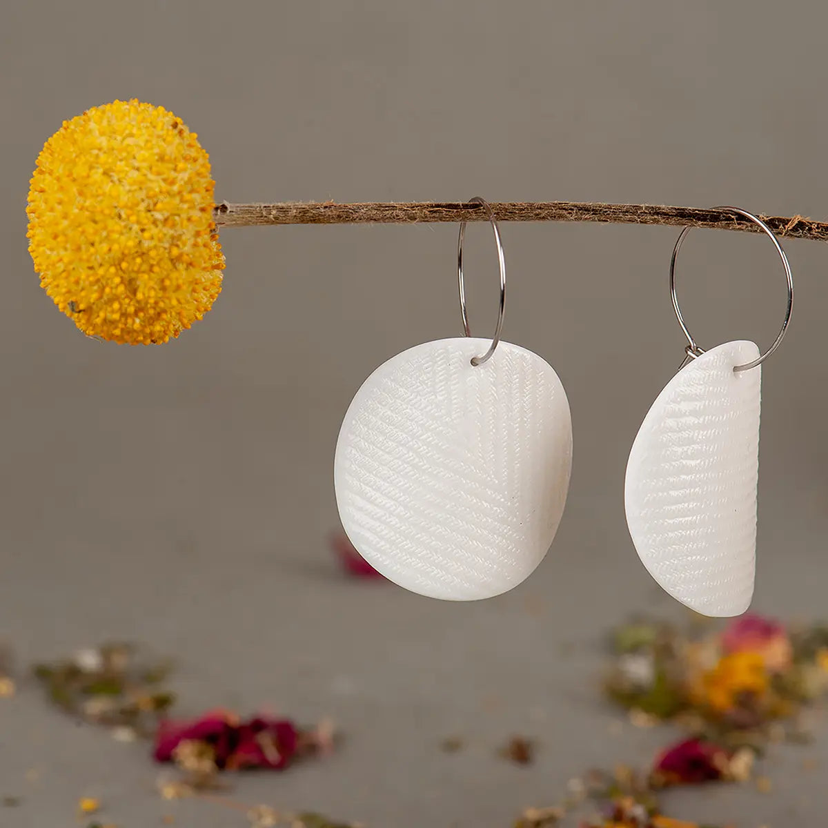 Two Urchic Porcelain "Moon" earrings in white geometric shapes hang from a thin branch with a yellow dried flower. Scattered petals are seen on the surface below against a neutral background, perfectly embodying the essence of minimalist accessories.