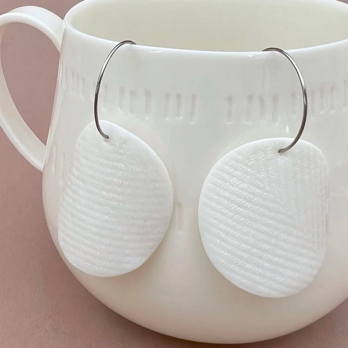 White ceramic mug with minimalist accessories, showcasing Urchic Porcelain's textured handmade "Porcelain Earrings - Moon" hanging on the rim against a light brown background.