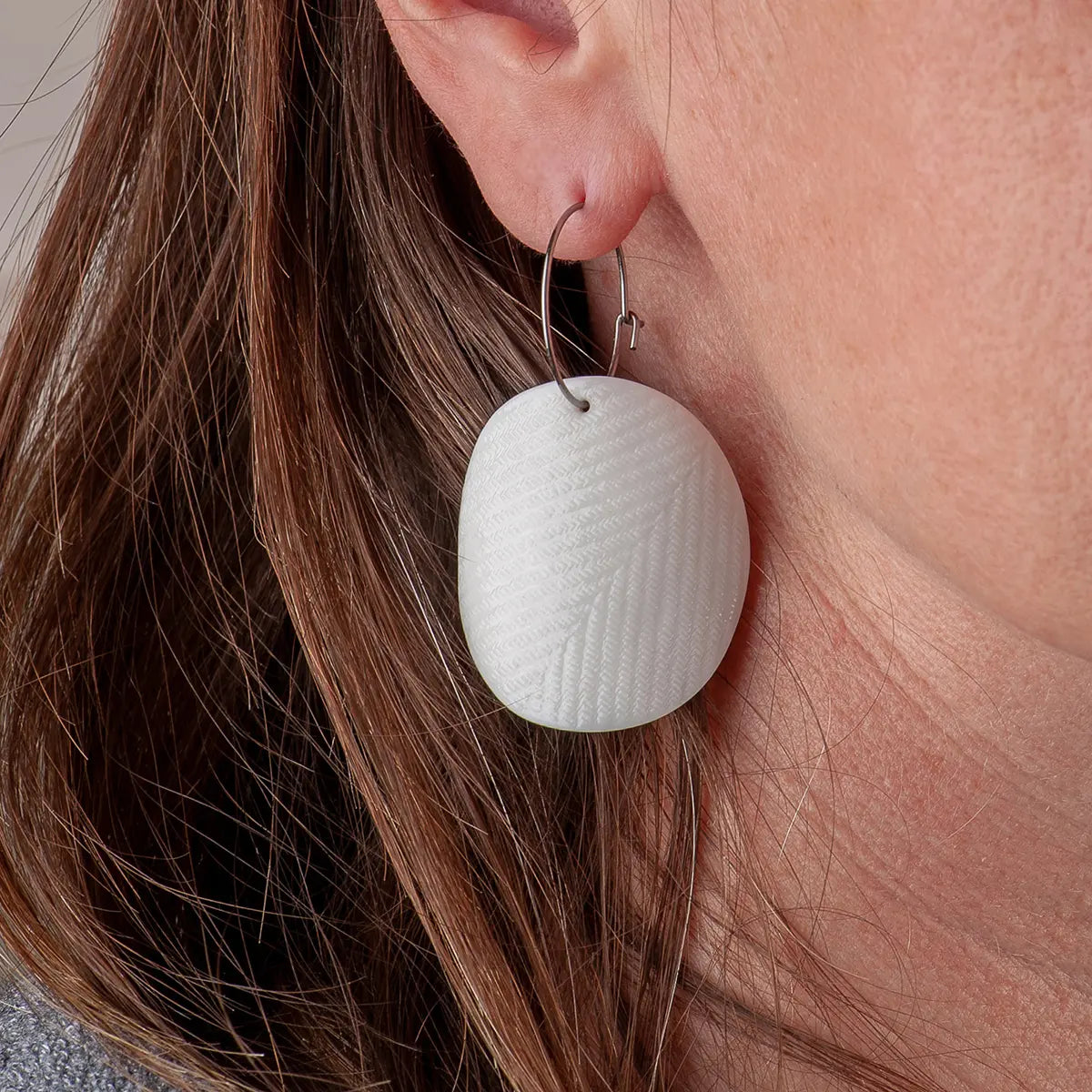 Close-up of a woman's ear wearing Urchic Porcelain's hypoallergenic surgical steel hoops adorned with a large, textured, oval-shaped white pendant from their Porcelain Earrings - Moon collection, showcasing minimalist accessories.