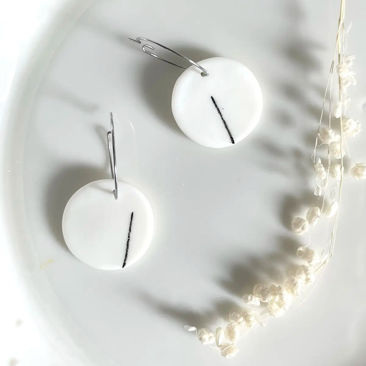 Two white round Porcelain Earrings - One by Urchic Porcelain, each featuring a single black line, are displayed on a white surface next to a sprig of small white flowers. Their minimalist design exudes elegance, making them perfect for any occasion.