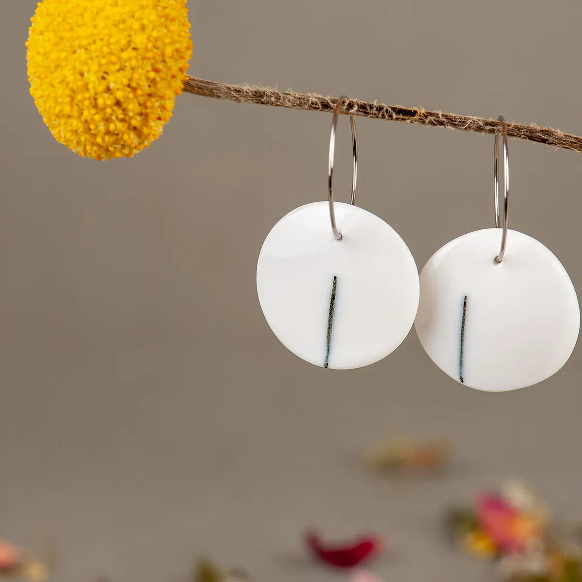 A pair of "Porcelain Earrings - One" by Urchic Porcelain, featuring minimalist black lines on round porcelain beads, hang from a twig next to a yellow flower against a neutral background, showcasing their hypoallergenic hoops designed for sensitive ears.