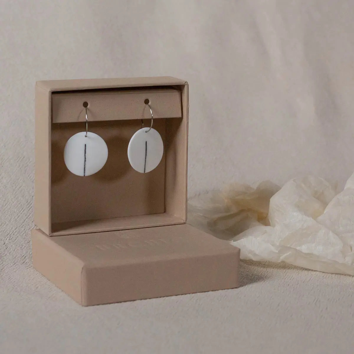 A pair of Urchic Porcelain's "Porcelain Earrings - One" featuring a vertical line design in the center are elegantly displayed in an open beige jewelry box against a neutral background, highlighting their minimalist aesthetic.