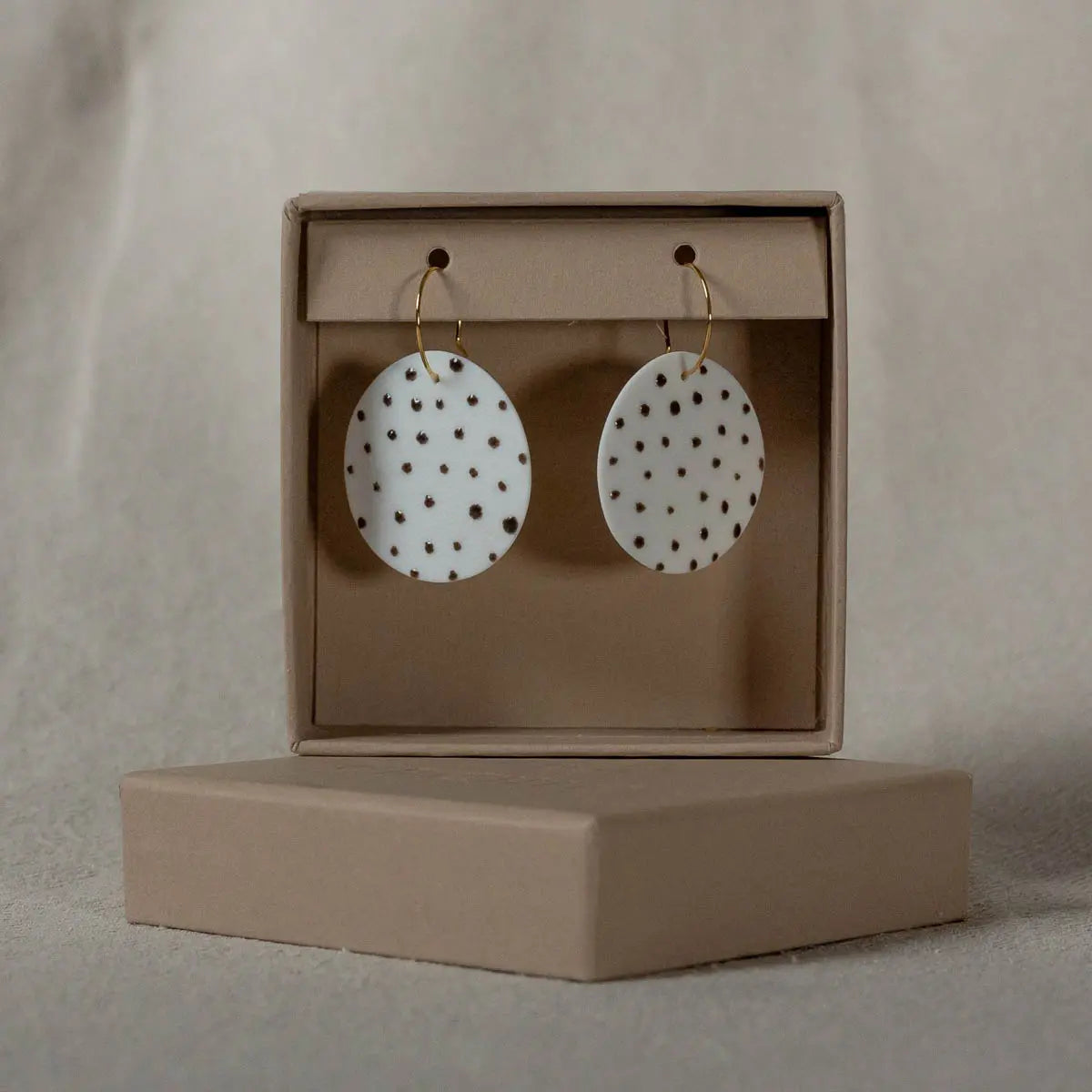 A pair of Porcelain Earrings - Polka dots from Urchic Porcelain, featuring handcrafted white oval porcelain with small brown spots, displayed in a beige cardboard box with a lid.