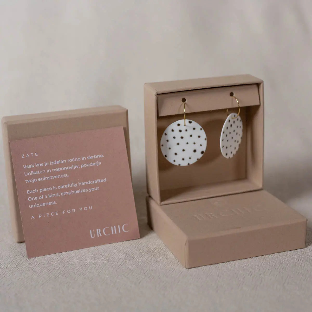 A pair of round, white porcelain earrings with black polka dots is displayed in an open beige jewelry box. A card with the product description stands beside it. The handcrafted earrings are branded as "Urchic Porcelain" and are hypoallergenic for sensitive ears.