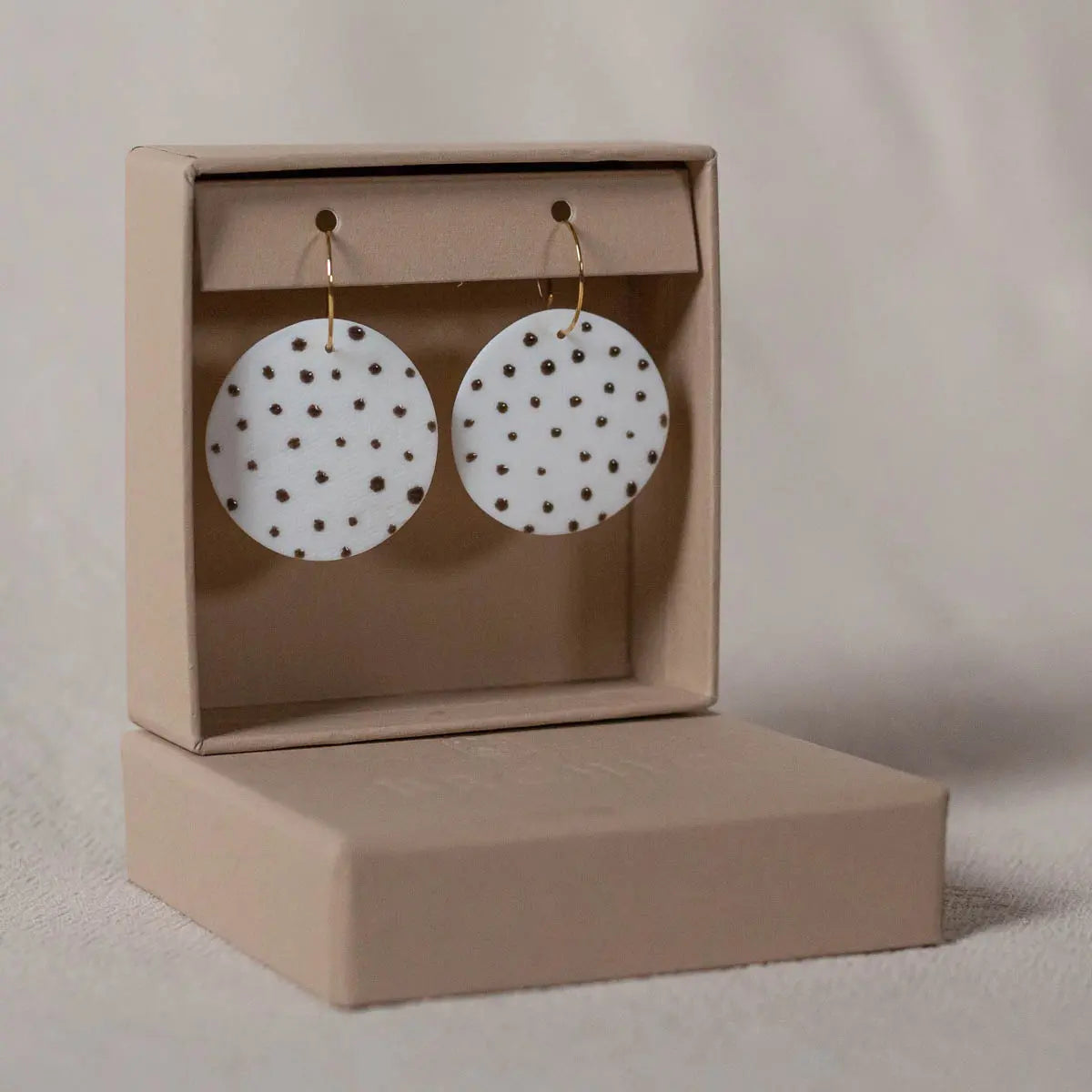 A pair of Porcelain Earrings - Polka dots by Urchic Porcelain, featuring round white handcrafted porcelain with black dot patterns, are displayed in an open beige box on a beige surface.