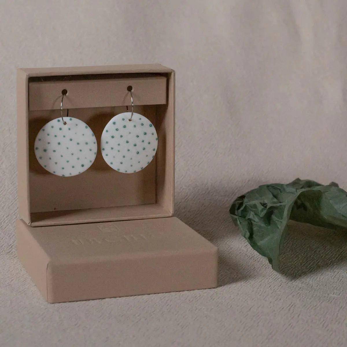 A pair of Urchic Porcelain's "Porcelain Earrings - Polka dots," featuring white round porcelain with green dots, meticulously handcrafted and hypoallergenic, displayed on a beige cardboard box next to a green leaf on a textured fabric background.