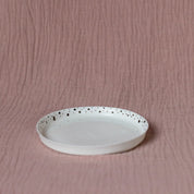 A Mini serving plate with black dots from Urchic Porcelain, adorned with elegant black dots along the rim, is placed on a textured light pink fabric background.