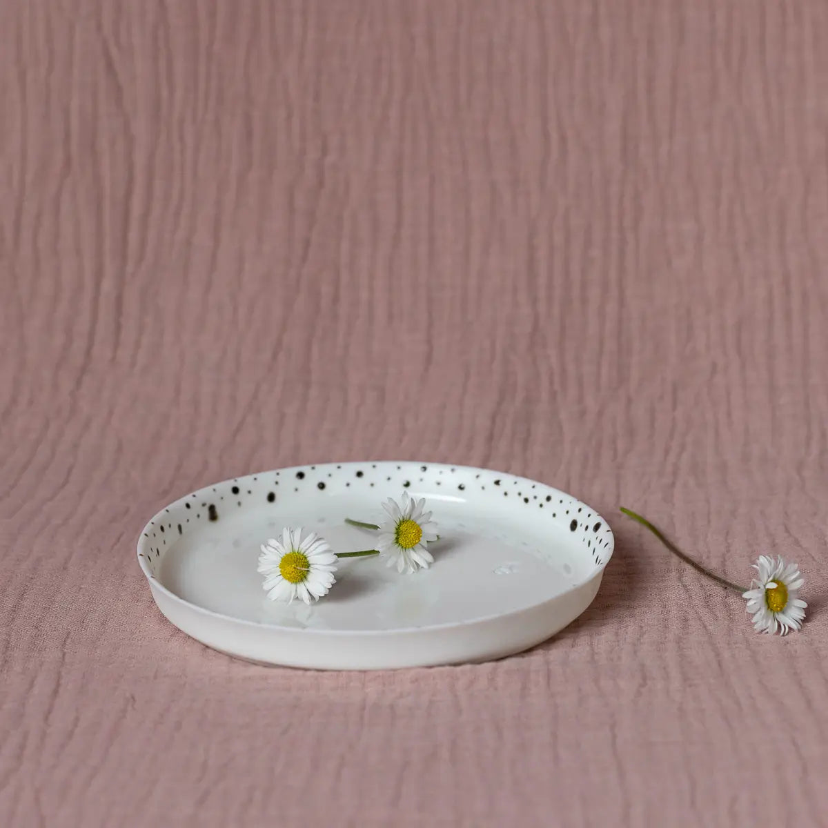 A handmade Mini serving plate with elegant black dots from Urchic Porcelain features two daisies set against a textured pink background, while a third daisy lies beside the plate.