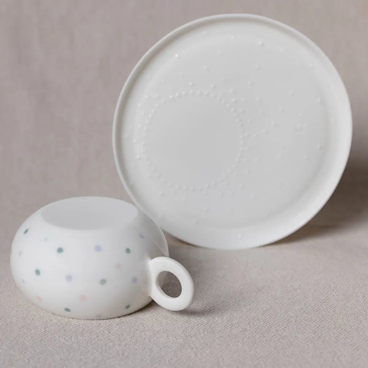 A white ceramic Nova Cup by Urchic Porcelain, adorned with small pastel polka dots, lies upside down next to a matching Saucer for Nova Cup on a beige surface.