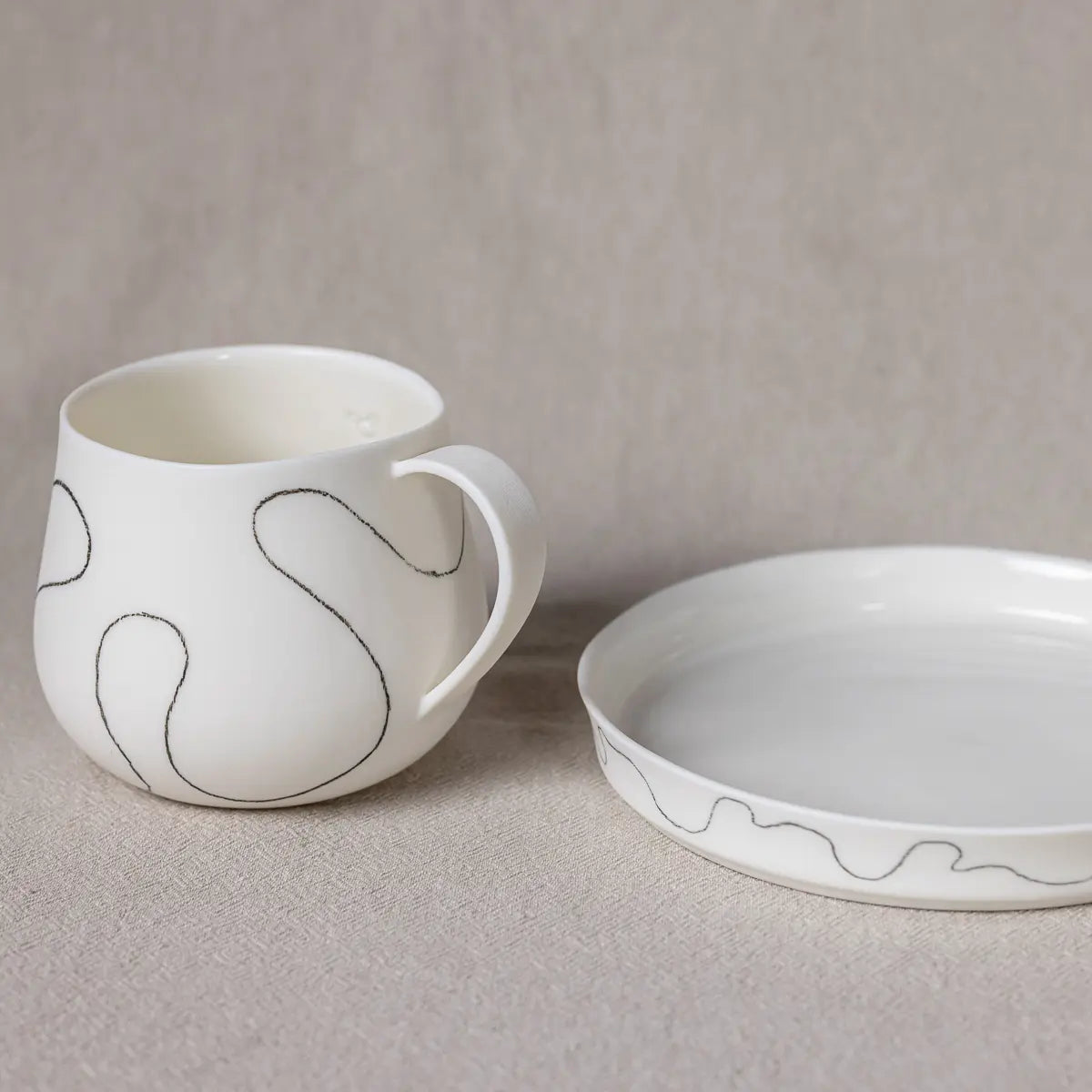 A white ceramic Dolce dessert serving plate with a rim from Urchic Porcelain, adorned with abstract black line patterns, sits on a beige surface. The matching cup has a curved handle and features similar line designs, resembling fine porcelain breakfast dishes.