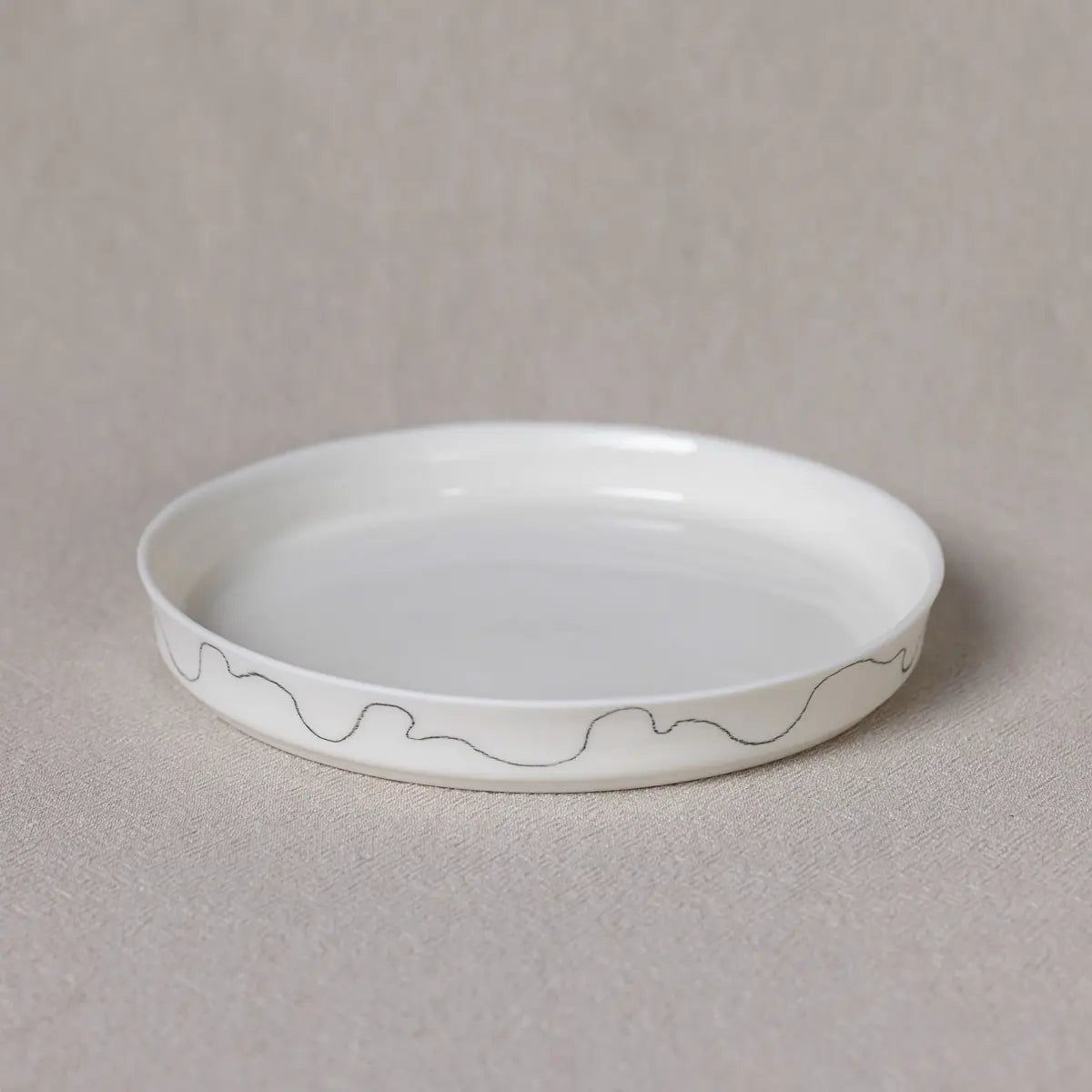 A Dolce dessert serving plate with a simple black wavy line design around its rim by Urchic Porcelain, placed on a beige surface.