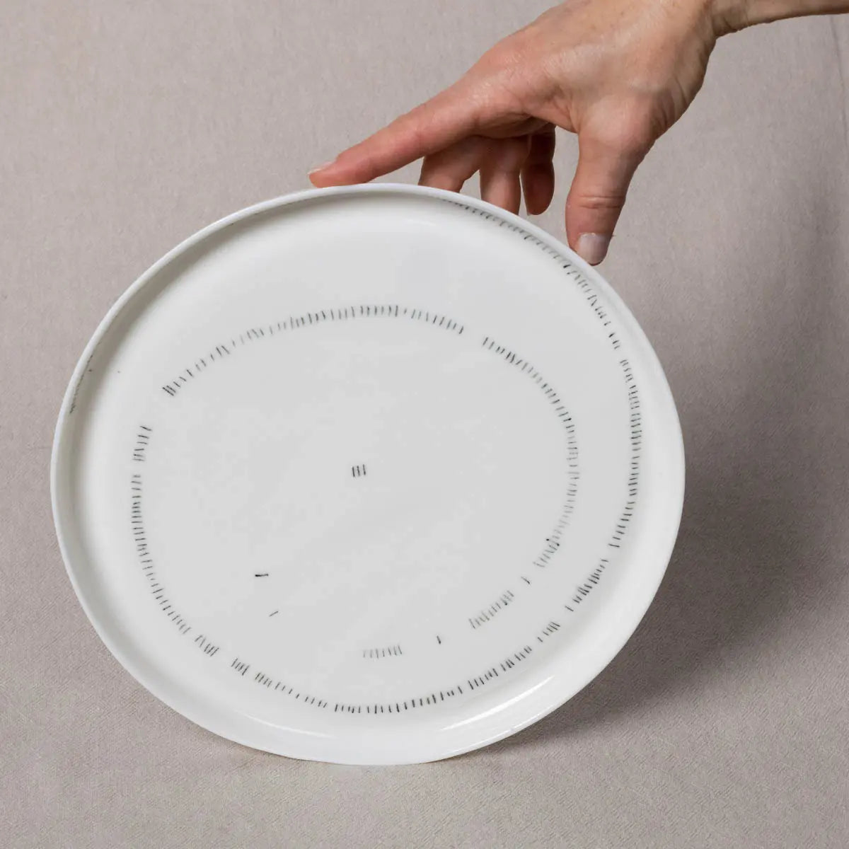 A hand gently holds the edge of an Urchic Porcelain TRDINKA dinner plate with rim featuring a minimalist Corn fields pattern.