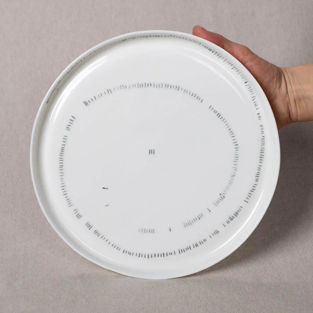 A hand holds a TRDINKA dinner plate with rim, adorned with a Corn fields pattern from Urchic Porcelain. The background is a light beige fabric, subtly enhancing the elegance of this handcrafted dinnerware.