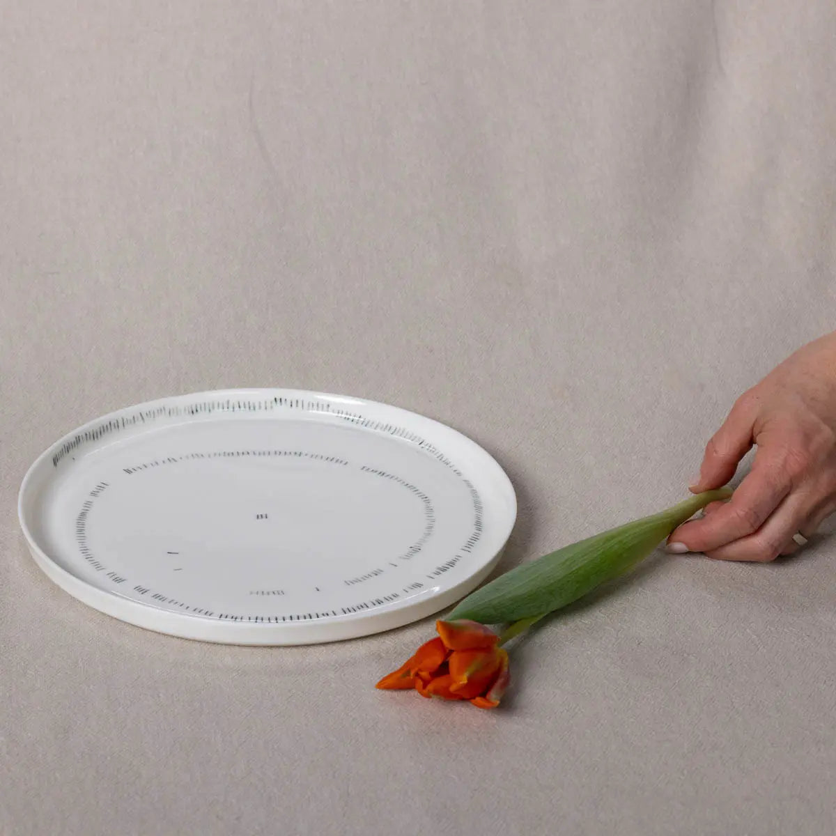 A hand holds an orange tulip by its stem next to a TRDINKA dinner plate with a Corn fields pattern by Urchic Porcelain on a beige surface.