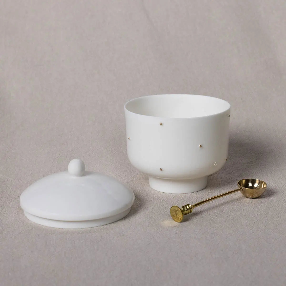 A white Urchic Porcelain Plečnik Sugar Bowl with gold accents sits next to a small brass spoon, all elegantly placed on a neutral-colored surface.