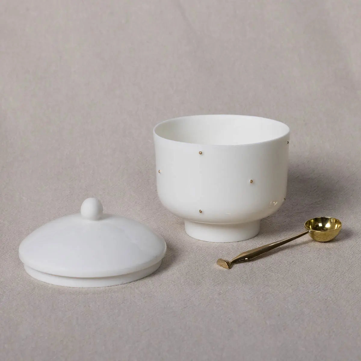 A white Urchic Porcelain Plečnik Sugar Bowl & Spoon Set, featuring a lid and a small brass spoon, placed on a light beige surface.