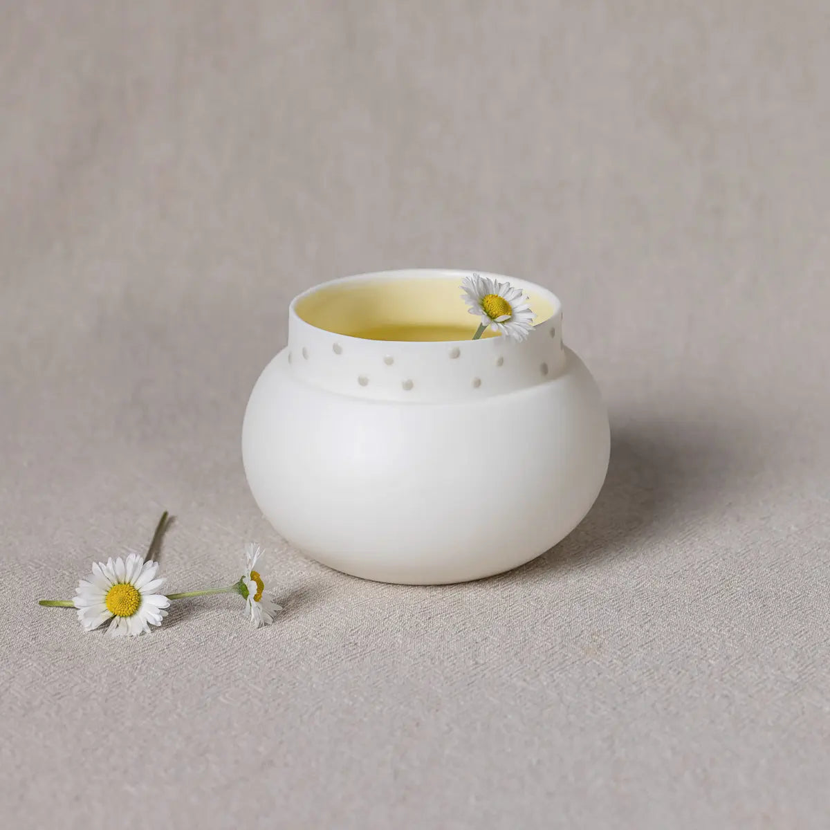 The Vase Dama by Urchic Porcelain, a small white ceramic vase with a yellow interior and dotted design, holds a single daisy flower as it sits on a neutral fabric background. This charming decorative element is accompanied by two additional daisies placed beside the vase.