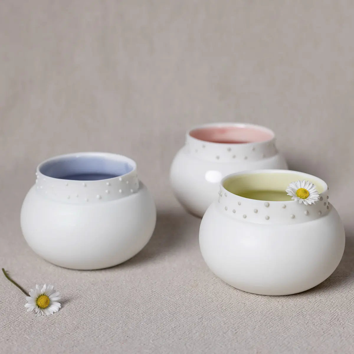 Three Vase Dama pieces by Urchic Porcelain, featuring white ceramic exteriors with blue, pink, and yellow interiors, are elegantly arranged on a beige surface. Each vase is adorned with a daisy flower, adding a charming decorative element to the scene.