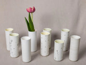 A group of eight white ceramic Vases Kiki, some adorned with delicate black blossom patterns from Urchic Porcelain, arranged on a light beige background. One elegant vertical vase holds two pink tulips with green leaves, serving as a decorative accent.