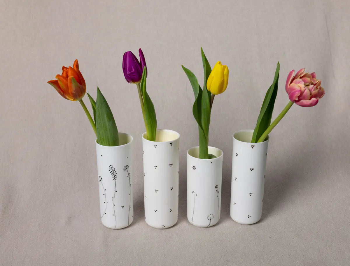 Four tulips in yellow, purple, red, and pink are arranged in four elegant vertical vases featuring the minimalistic black Blossoms Pattern on translucent porcelains by Urchic Porcelain's Vase Kiki collection, set against a beige background.