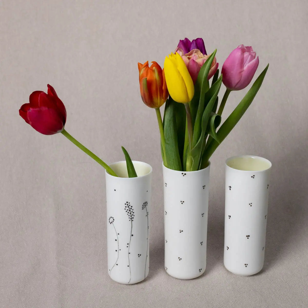 Three elegant Vase Kikis from Urchic Porcelain, each made of translucent porcelain adorned with the Blossoms Pattern in black dots, hold different colored tulips—red, orange, yellow, and purple. Arranged beautifully against a plain beige background, this decorative accent adds a touch of sophistication to any space.