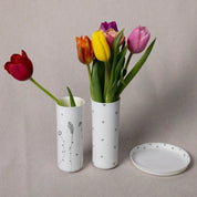 Two white, translucent porcelain vases identified as "Vase Kiki, Blossoms Pattern" by Urchic Porcelain, each holding tulips, accompanied by a matching white plate, all set against a neutral background. These elegant vertical vases serve as the perfect decorative accent.