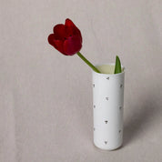 A single red tulip in the Vase Kiki featuring a Blossoms Pattern by Urchic Porcelain, made of translucent porcelain with small black dot patterns, sits gracefully on a light gray fabric background.