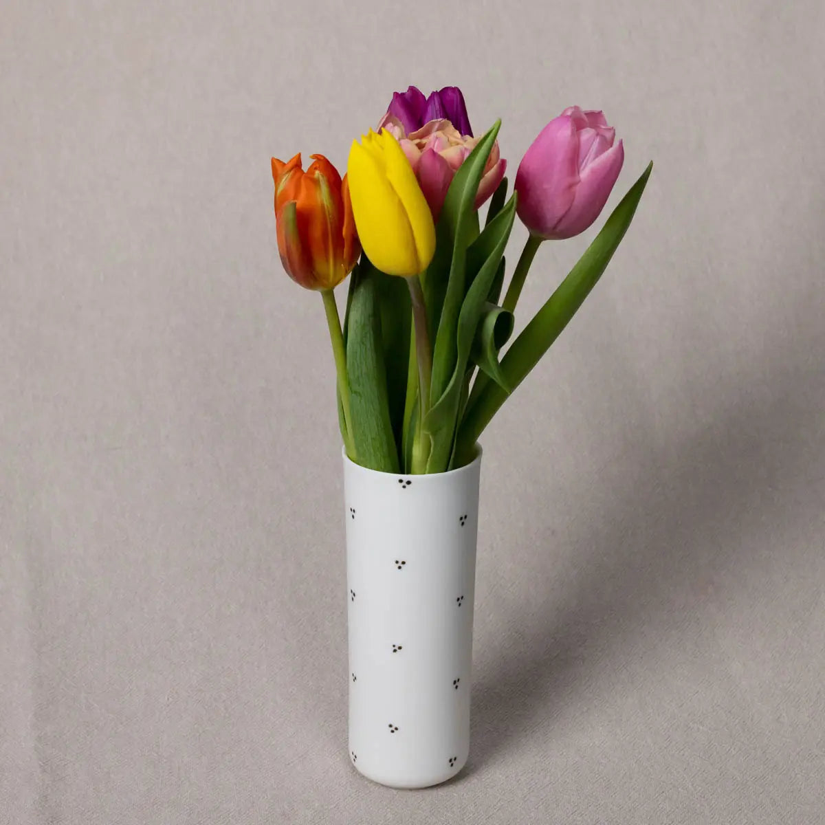 Vase Kiki, Blossoms Pattern from Urchic Porcelain is a small vertical vase made of translucent porcelain with elegant black speckles, holding five tulips in various colors—orange, yellow, purple, and pink—against a neutral background. This decorative accent adds a touch of sophistication to any space.