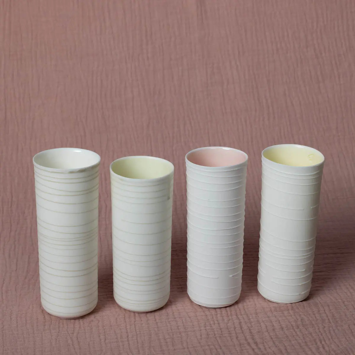 Four stackable, cylindrical ceramic cups from Urchic Porcelain's "Vase Kiki" series, featuring a lines pattern and varying interior colors of white, yellow, pink, and yellow, are arranged on a soft pink textured surface. These stylish cups serve as a charming decorative accent for any setting.