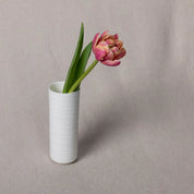 A single pink flower with green leaves stands in the elegant Vase Kiki with a lines pattern from Urchic Porcelain, placed on a beige surface as a beautiful decorative accent.