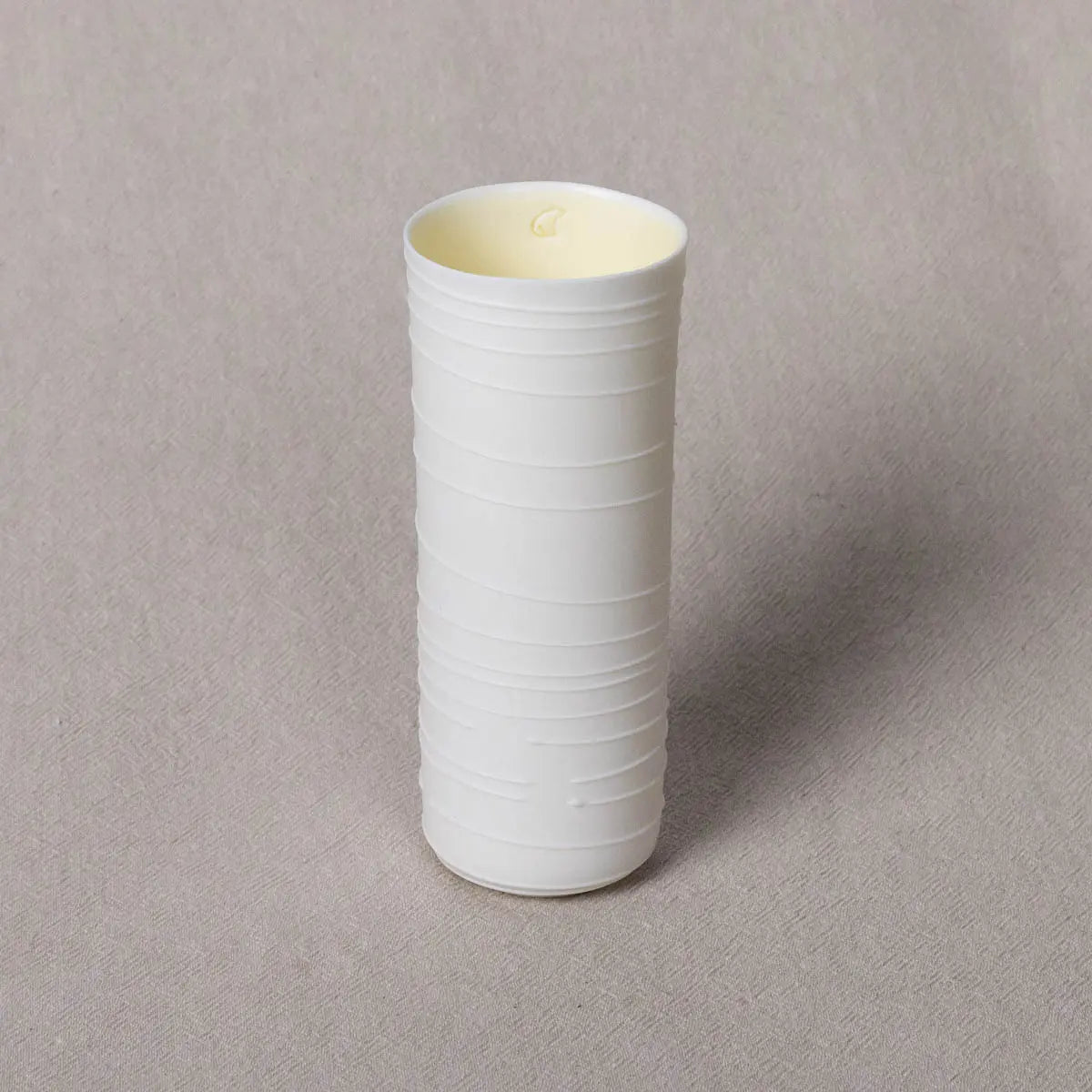 The Vase Kiki from Urchic Porcelain is an elegant cylindrical, white ceramic vase with a textured lines pattern and an open top, standing gracefully on a gray surface and serving as a sophisticated decorative accent.