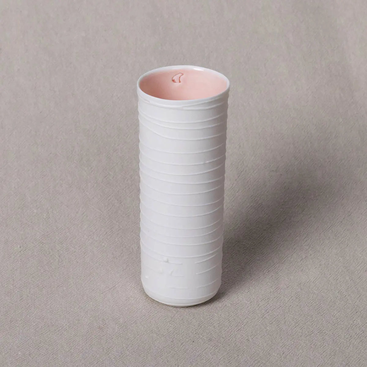 The Vase Kiki from Urchic Porcelain, featuring a cylindrical shape with white ceramic adorned in horizontal ridges and a light pink interior, stands on a beige textured surface. This elegant vase makes for a sophisticated decorative accent in any room.