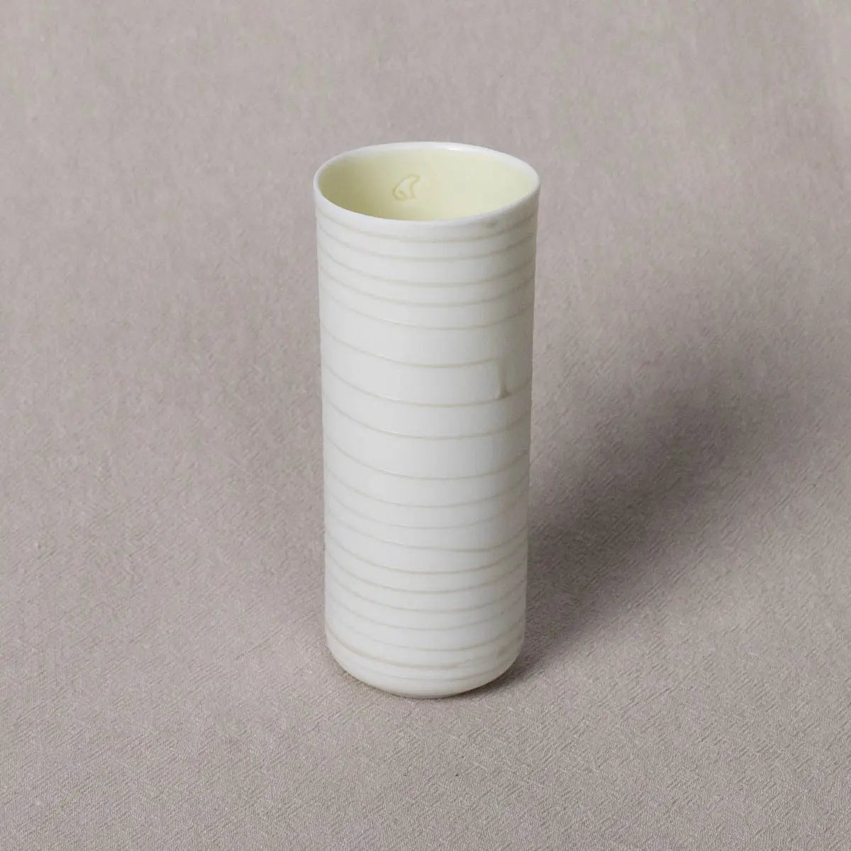 A tall, white ceramic Vase Kiki with subtle horizontal lines pattern from Urchic Porcelain stands on a beige surface, offering a decorative accent reminiscent of translucent porcelain.