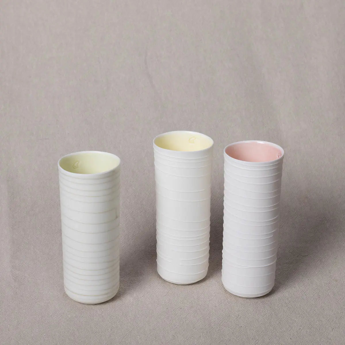 Three vertically ribbed cylindrical *Vase Kiki* in white porcelain from *Urchic Porcelain*, with interiors colored yellow, pastel yellow, and pink, are placed on a beige surface. These elegant vessels serve as both functional pieces and decorative accents.