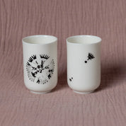 The Soulmates Set of Two Cups by Urchic Porcelain showcases the elegant Vurnik Carnation Pattern, featuring two white ceramic espresso cups with black abstract designs. One cup displays a detailed central pattern while the other features small scattered elements, all set against a textured pink backdrop.