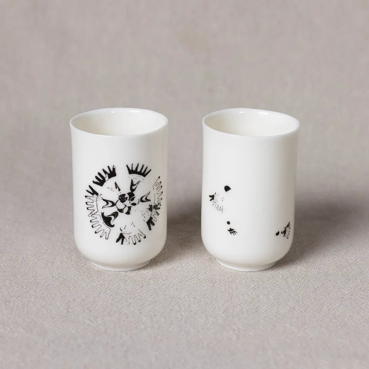 The Soulmates Set of Two Cups by Urchic Porcelain, part of the Vurnik Carnation Pattern collection, includes two white porcelain espresso cups. One cup displays a detailed floral design while the other features sparse, abstract patterns. Both cups are elegantly arranged on a neutral fabric background.