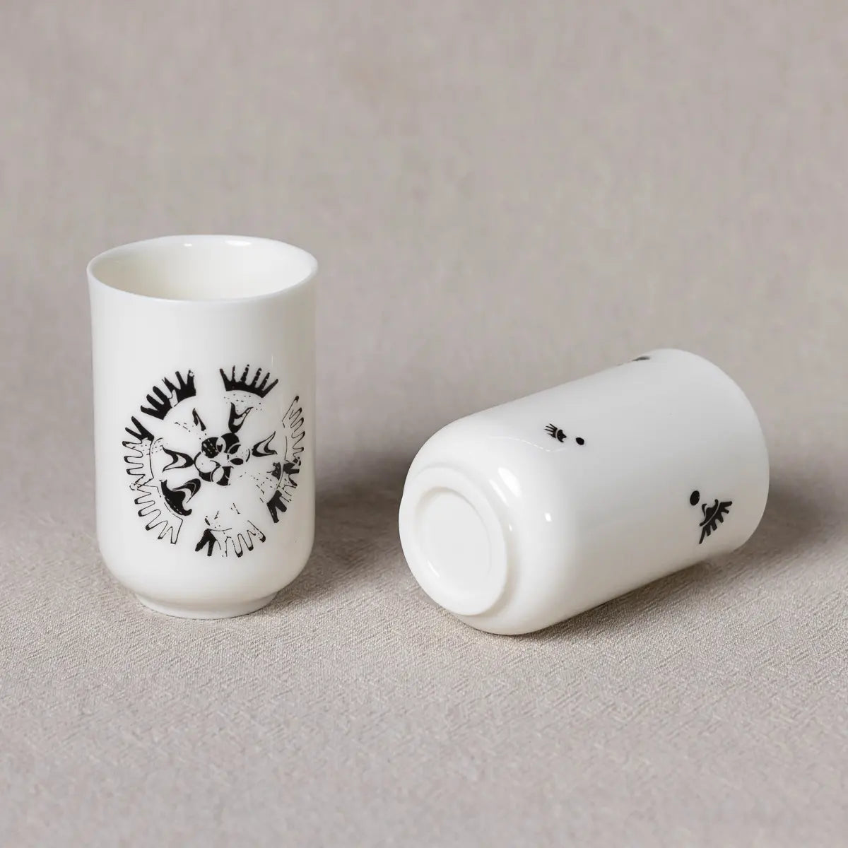 Two white espresso cups from the Soulmates Set of Two Cups, Vurnik Carnation Pattern by Urchic Porcelain: one stands upright, displaying a black abstract design; the other lies on its side, showing a partial design. Both are placed on a textured beige surface.