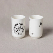 Two white porcelain cups from Urchic Porcelain's Soulmates Set of Two Cups, Vurnik Carnation Pattern; one cup features a large central black abstract design, while the other has small scattered black designs. Both are part of the Vurnik collection and are set against a plain beige background.