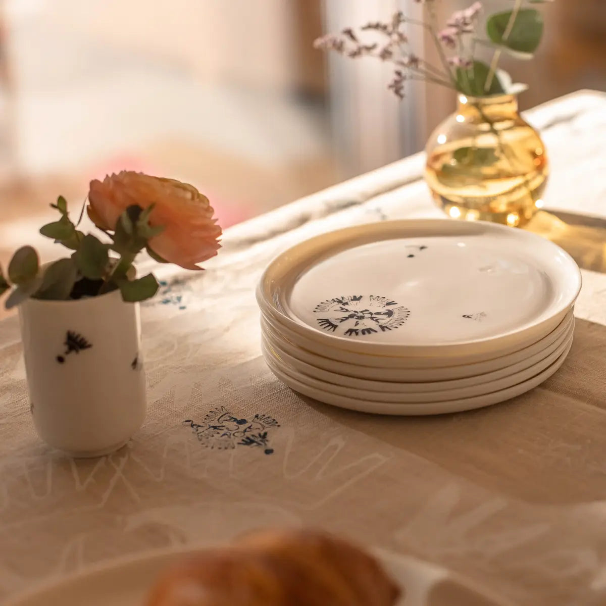 A stack of ceramic plates with floral designs sits on a table next to a small vase containing a pink flower, an espresso cup from the Soulmates Set of Two Cups in Vurnik Carnation Pattern by Urchic Porcelain, and a golden round vase with a sprig, all arranged on a patterned tablecloth.