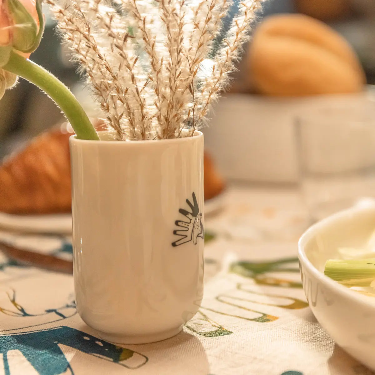 A white ceramic vase with dried grass stems and a green plant on a patterned tablecloth, featuring an elegant porcelain cup from the Soulmates Set of Two Cups, Vurnik Carnation Pattern by Urchic Porcelain in the background beside blurred plates and bread.