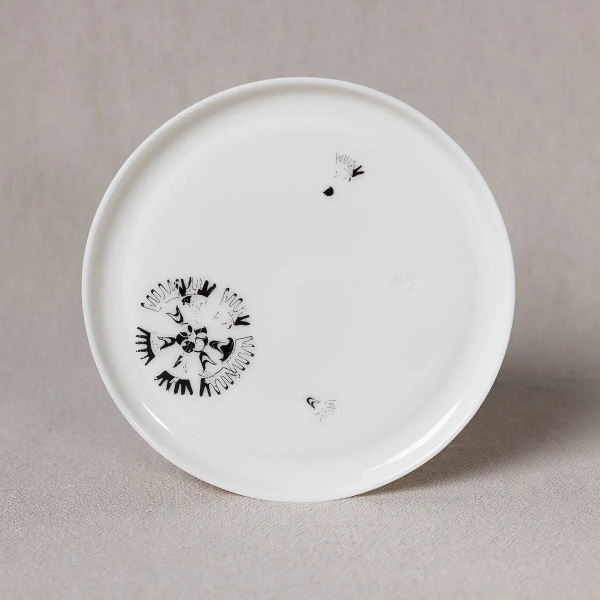 A Vurnik Carnation pattern plate from Urchic Porcelain, featuring minimal black abstract designs near the rim and a clean, white circular surface, is placed on a light grey fabric.