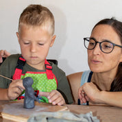 At GLINARIJE NA LONTRGU: Poletna mala šola gline za otroke – AKONTACIJA by Urchic Porcelain, a boy shapes clay as a woman with glasses guides him, nurturing children's creativity at the pottery table.