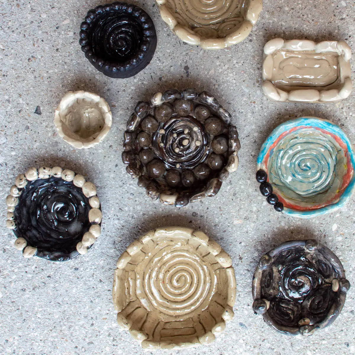 Eight vibrant, handmade clay bowls and dishes by children at the GLINARIJE NA LONTRGU: Poletna mala šola gline za otroke - AKONTACIJA (Urchic Porcelain) display coiled and beaded designs on a textured concrete surface.
