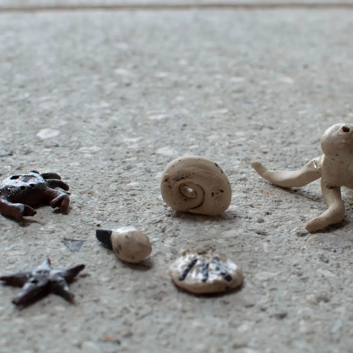 Six small sea creature and shell sculptures, created by children at Urchic Porcelain’s "GLINARIJE NA LONTRGU: Poletna mala šola gline za otroke - AKONTACIJA" workshop, are displayed on a speckled, light surface.