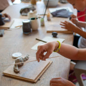 At GLINARIJE NA LONTRGU: Poletna mala šola gline za otroke by Urchic Porcelain, children shape objects with clay and wooden tools, building creativity and fine motor skills.