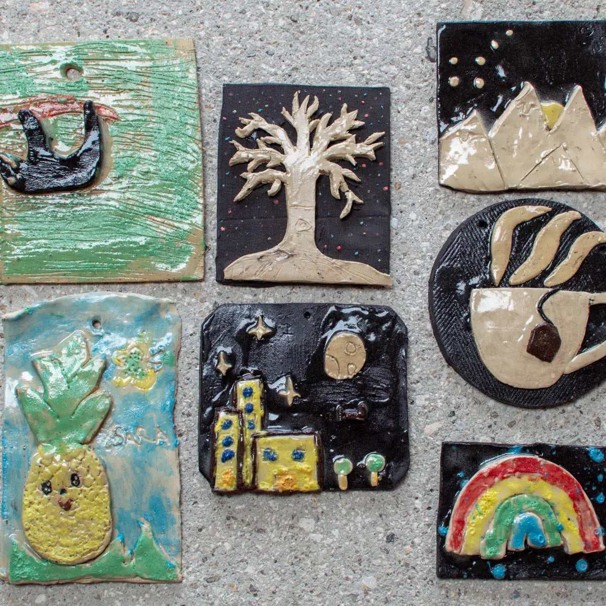 Seven ceramic tiles featuring designs like a bird, tree, mountains, pineapple, cityscape, coffee cup, and rainbow by Urchic Porcelain—ideal for sparking creativity at GLINARIJE NA LONTRGU: Poletna mala šola gline za otroke - AKONTACIJA.