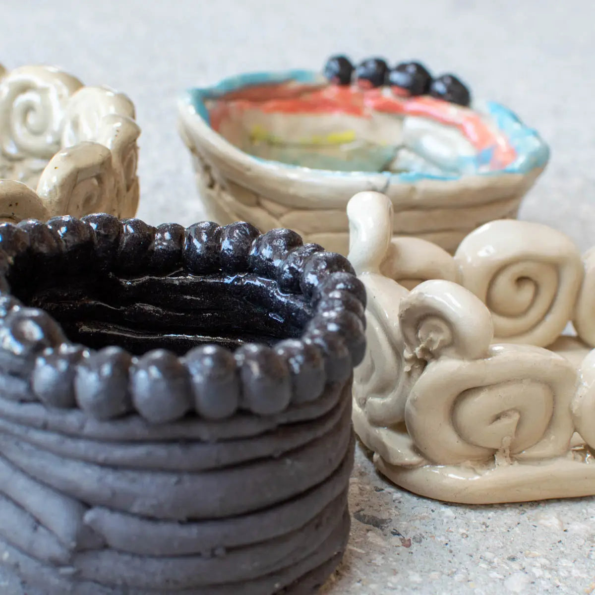 Four unique, hand-built clay pots from Urchic Porcelain’s GLINARIJE NA LONTRGU: Poletna mala šola gline za otroke – AKONTACIJA display children's creativity and fine motor skills, crafted in a summer workshop and shown on a light surface.