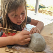 At Urchic Porcelain's GLINARIJE NA LONTRGU: Poletna mala šola gline za otroke – AKONTACIJA, children use wooden tools to shape clay on boards, boosting creativity and fine motor skills at a fun summer pottery workshop.