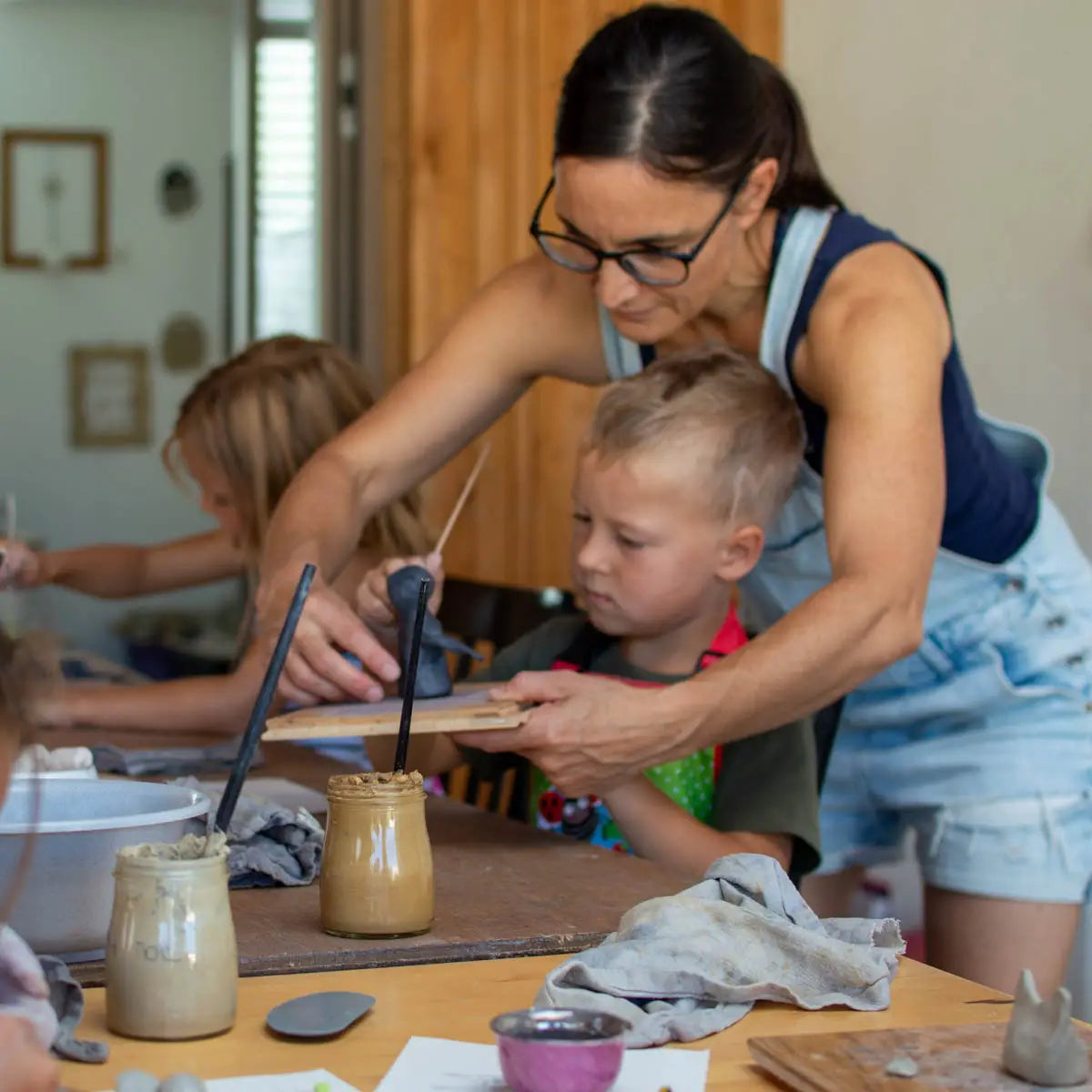 At GLINARIJE NA LONTRGU: Poletna mala šola gline za otroke - AKONTACIJA by Urchic Porcelain, an adult guides a child shaping clay, fostering fine motor skills while other kids work on creative pottery in the background.
