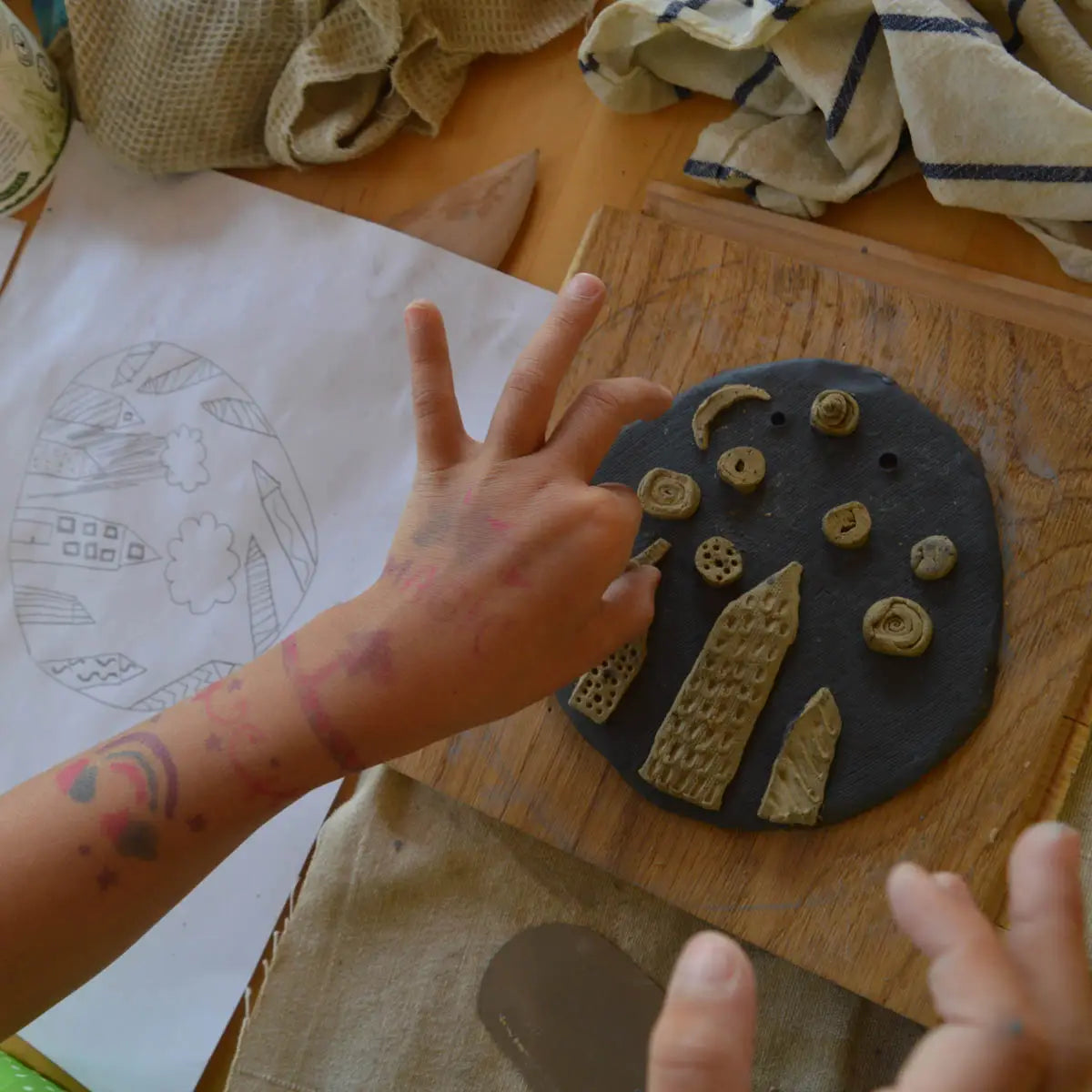 At Urchic Porcelain's "GLINARIJE NA LONTRGU: Poletna mala šola gline za otroke – AKONTACIJA," children build creativity by decorating clay pieces with sculpted shapes, drawings, and fabric at a fun summer pottery workshop.